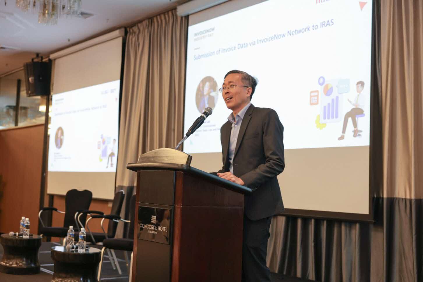 Mr Dennis Lui, IRAS’ Deputy Commissioner (Indirect Taxes, Law & Investigation Group), stands on the stage behind a desk with microphones, addressing a gathering of specialists and journalists, and announces the GST InvoiceNow Requirement during the IMDA’s InvoiceNow Industry Day