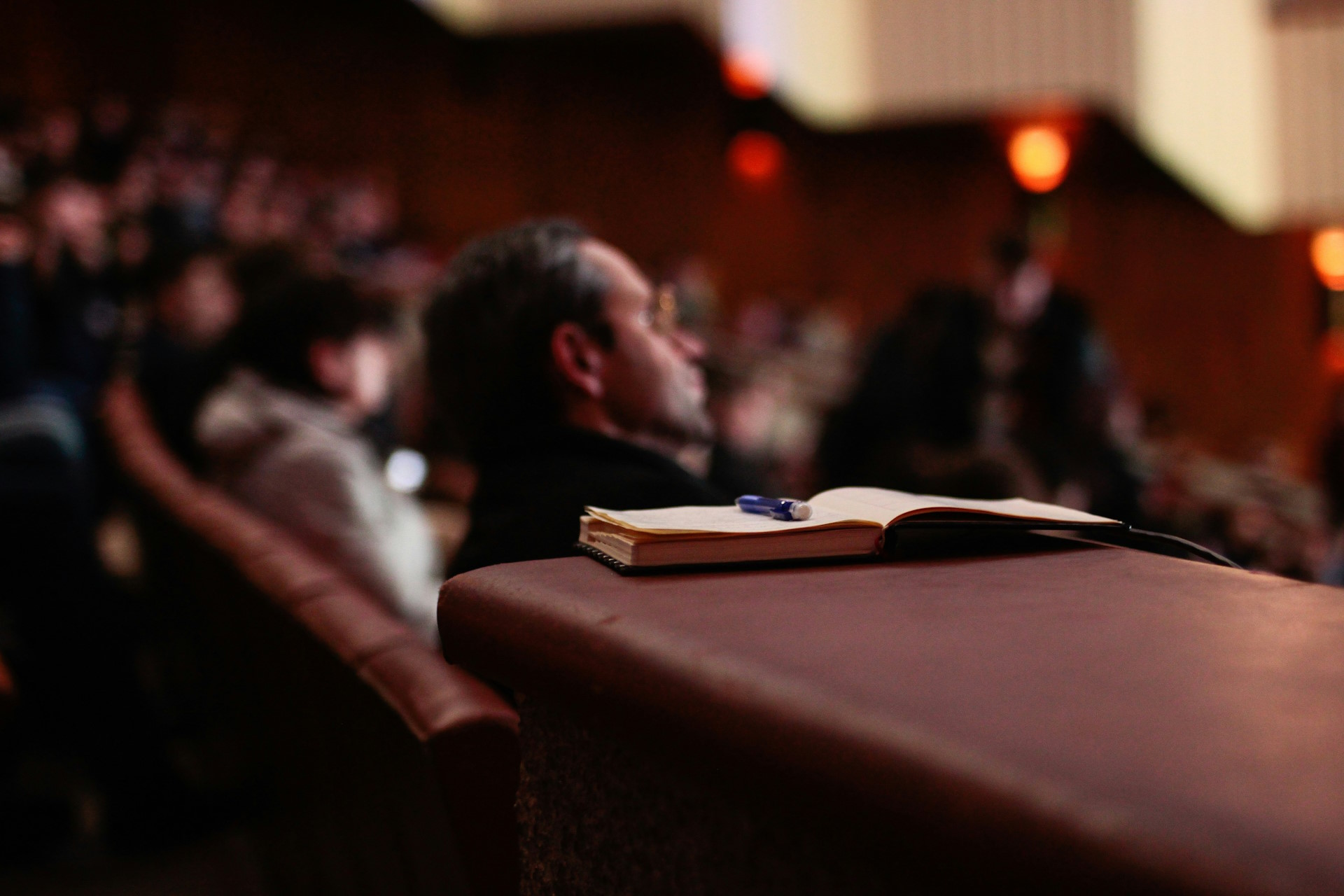 Focus on a paper notebook with conference attendees sitting blurred in the background